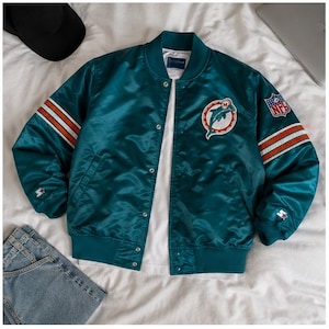 May include: A teal satin bomber jacket featuring the Miami Dolphins logo on the left chest, an NFL shield patch on the left sleeve, and white and orange stripes on the sleeves. The jacket has a snap-button front closure and ribbed trim.