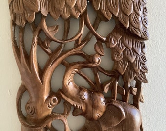 May include: A wooden wall hanging sculpture of two elephants standing under a tree with intricate carvings. The sculpture is made of dark brown wood and has a rustic, natural look.