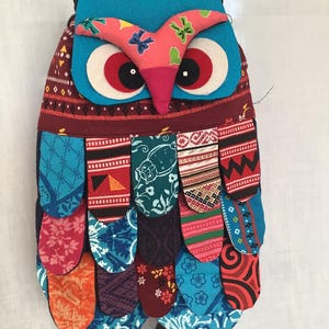 Puede incluir: Una colorida bolsa de patchwork con forma de búho con un cuerpo azul, ojos rojos y pico rosa. La bolsa está hecha de diferentes telas con varios patrones y colores, incluyendo rojo, azul, verde y naranja.