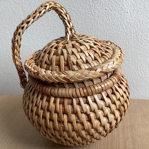 May include: A woven wicker basket with a lid and a handle. The basket is light brown and has a natural finish.