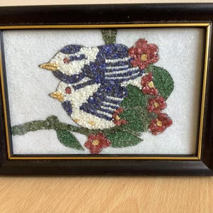 May include: Framed artwork featuring two birds perched on a branch, crafted from small, colorful stones. The birds have blue and white striped bodies, with red accents. Red flowers and green leaves surround the birds, set against a white background.