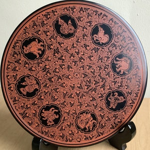 Puede incluir: Un plato redondo negro con un borde elevado, decorado con un intrincado patrón floral naranja y figuras de animales negros. El plato está sobre un soporte negro.