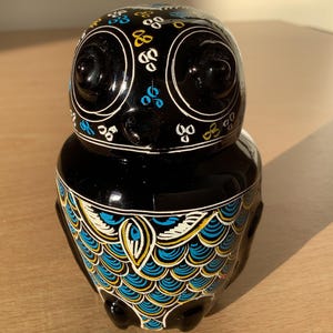 May include: Black owl-shaped ceramic jar with blue, yellow, and white painted details. The jar has a round body and a flat base.