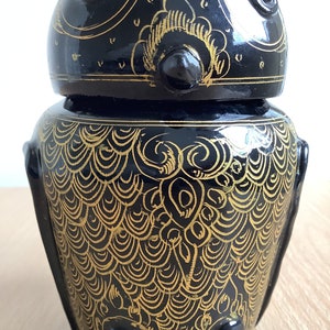 May include: Black and gold owl-shaped ceramic jar with intricate details. The jar has a round body and a lid with a raised owl face. The owl's eyes are black and the body is covered in a gold pattern.