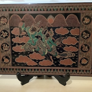 May include: A black wooden panel with a gold and green painted scene of two figures flying through the clouds. The panel is framed with a decorative border and is displayed on a black stand.