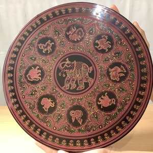 Könnte beinhalten: Ein rundes, schwarzes Tablett mit einem rosa und goldenen Design. Das Tablett zeigt einen zentralen Elefanten, umgeben von kleineren Elefanten und floralen Mustern.