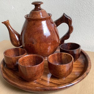May include: A handcrafted wooden tea set with a teapot, lid, four cups, and a serving tray. The set is made of polished, dark brown wood. The teapot has a curved spout and handle. The cups are small and round. The tray is oval-shaped.