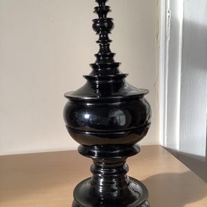 May include: A black decorative urn with a glossy finish. The urn has a bulbous midsection, a tiered base, and a slender, pointed top. The design features multiple stacked elements, creating a visually interesting silhouette. The urn is placed on a light-coloured wooden surface.