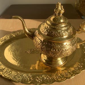 May include: A golden tea set with intricate floral patterns. The set includes a teapot with a decorative handle and lid, and a matching oval tray. The teapot sits on a pedestal base. The set is made of a shiny, reflective metal.