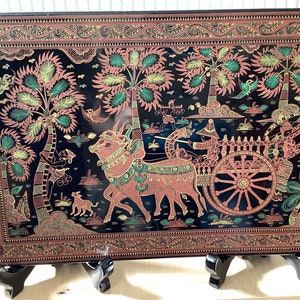 May include: A black lacquered wooden panel with a gold and red painted scene of a bullock cart with two oxen pulling it. The cart is decorated with gold and red details. The scene is set in a forest with palm trees and birds. The panel is framed with a gold and red border.