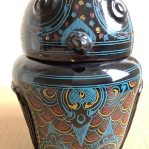 May include: Black ceramic owl-shaped jar with blue, red, and yellow painted designs. The jar has a lid and is decorated with intricate patterns.