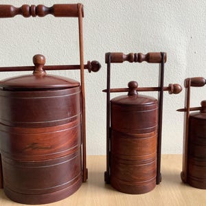 Könnte beinhalten: Drei mehrstöckige Holz-Essensbehälter in verschiedenen Größen. Jeder Behälter hat eine zylindrische Form, einen Deckel und einen Griff. Das Holz hat eine satte, dunkelbraune Farbe. Die Behälter stehen auf einer hellbraunen Oberfläche.