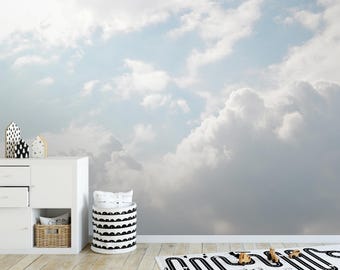 Puede incluir: Una cómoda blanca con dos cajones y un estante, una papelera de almacenamiento en blanco y negro con un patrón ondulado y un coche de juguete de madera sobre una alfombra con estampado blanco y negro. El fondo es un mural de pared de un cielo nublado.