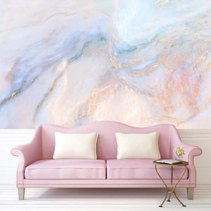 Puede incluir: Un sofá rosa con dos cojines blancos se encuentra frente a una pared con un diseño de mármol. La pared de mármol presenta remolinos de azul, rosa y dorado. Una pequeña mesa redonda con marco dorado y flores está junto al sofá.