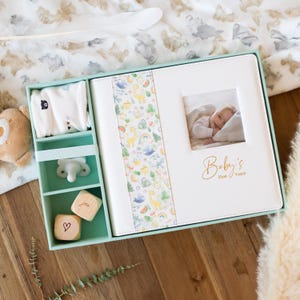May include: A white baby photo album with a green and white patterned spine. The album has a gold inscription that reads "Baby's First Years". The album is in a mint green box with a white pacifier, wooden blocks, and a white onesie.