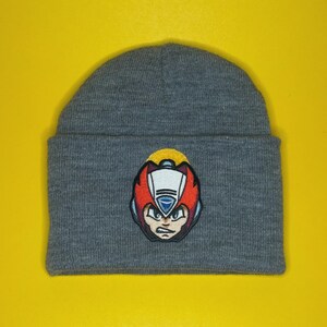 May include: A gray beanie featuring an embroidered patch of a video game character with a red helmet, white face, and blue accents. The beanie is made of a soft, knitted material and is suitable for cold weather. The background is a solid yellow.
