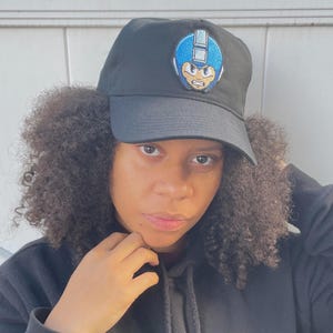 May include: Black baseball cap featuring a blue and yellow cartoon character patch. The cap has a curved brim and a classic six-panel design. The patch depicts a character with a blue helmet and a determined expression. This cap is a stylish accessory for fans of video games.