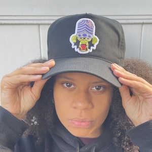 May include: Black baseball cap with a colorful embroidered patch featuring a cartoon character with a purple helmet and a green face. The cap has a curved brim and a structured fit.
