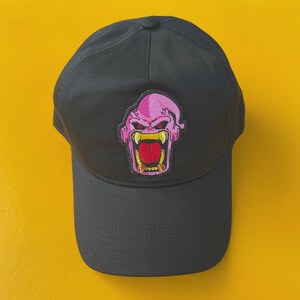 Pode incluir: Boné de beisebol preto com um patch de rosto de monstro rosa e vermelho. O patch tem um design detalhado com dentes afiados e uma expressão ameaçadora. O boné é feito de um tecido durável e tem uma aba curva.