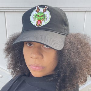 Pode incluir: Boné de beisebol preto com um patch de dragão verde e vermelho. O boné tem uma aba curva e uma coroa estruturada. O patch do dragão é circular e apresenta uma ilustração detalhada da cabeça de um dragão.