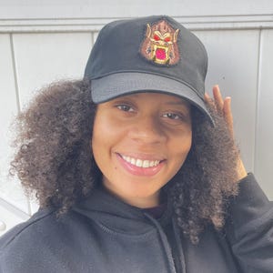 Pode incluir: Boné de beisebol preto com um patch bordado de uma criatura semelhante a um gorila com olhos vermelhos e boca dourada. O boné é feito de um tecido durável e tem uma aba curva.