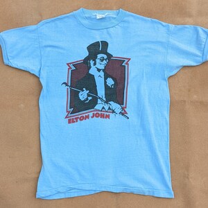 K&ouml;nnte beinhalten: Ein hellblaues T-Shirt mit einem Vintage-Motiv von Elton John, der einen Zylinder tr&auml;gt und einen Stock h&auml;lt. Das Motiv ist in Schwarzwei&szlig; mit roten Akzenten gehalten. Der Text "Elton John" ist unter dem Motiv gedruckt.