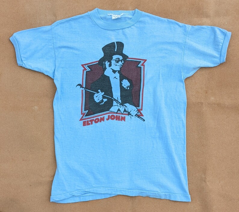 K&ouml;nnte beinhalten: Ein hellblaues T-Shirt mit einem Vintage-Motiv von Elton John, der einen Zylinder tr&auml;gt und einen Stock h&auml;lt. Das Motiv ist in Schwarzwei&szlig; mit roten Akzenten gehalten. Der Text "Elton John" ist unter dem Motiv gedruckt.