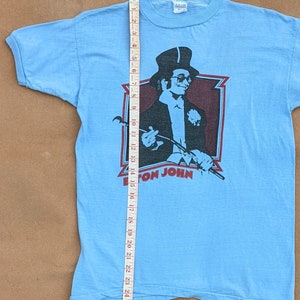 K&ouml;nnte beinhalten: Ein hellblaues T-Shirt mit einem Vintage-Elton-John-Motiv. Das Motiv zeigt ein Schwarzwei&szlig;bild von Elton John mit Zylinder und Sonnenbrille, der einen Stock h&auml;lt. Der Text "Elton John" ist in Rot unter dem Bild gedruckt.