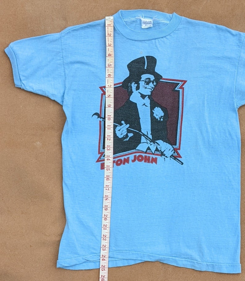 K&ouml;nnte beinhalten: Ein hellblaues T-Shirt mit einem Vintage-Elton-John-Motiv. Das Motiv zeigt ein Schwarzwei&szlig;bild von Elton John mit Zylinder und Sonnenbrille, der einen Stock h&auml;lt. Der Text "Elton John" ist in Rot unter dem Bild gedruckt.