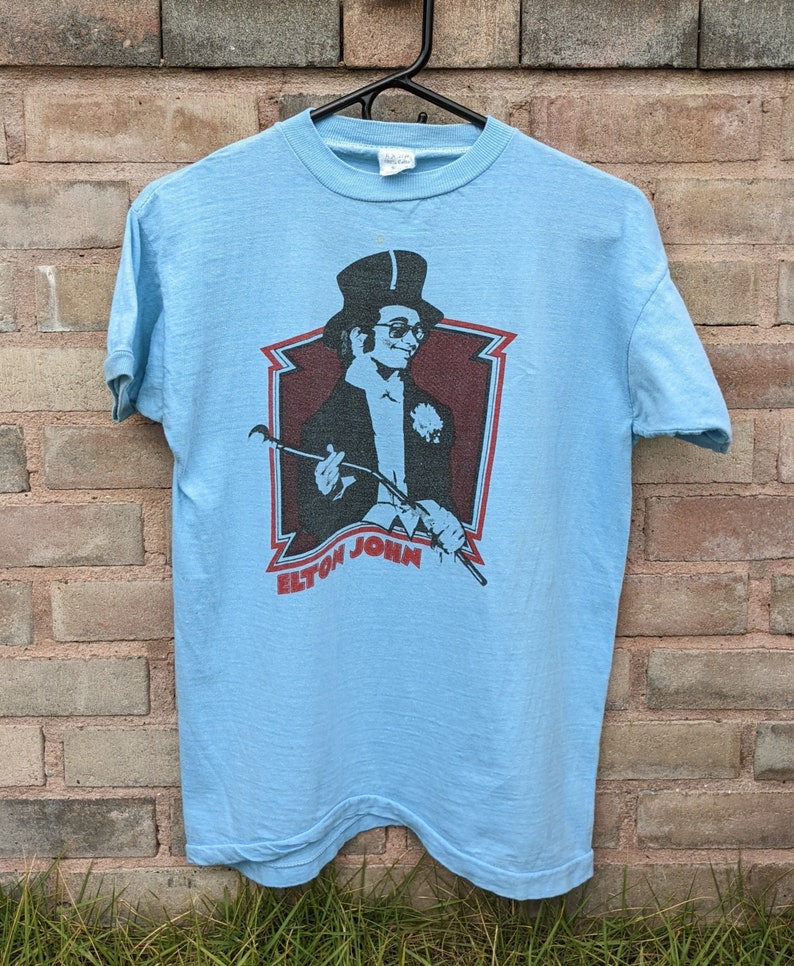 K&ouml;nnte beinhalten: Ein hellblaues T-Shirt mit einem Vintage-Motiv von Elton John, der einen Zylinder und eine Sonnenbrille tr&auml;gt. Das Motiv ist in einer roten und schwarzen Schildform mit dem Text "Elton John" darunter.