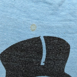 K&ouml;nnte beinhalten: Ein hellblaues T-Shirt mit einem Schwarzwei&szlig;bild einer Person mit Hut und Sonnenbrille. Das Bild ist leicht verblasst.