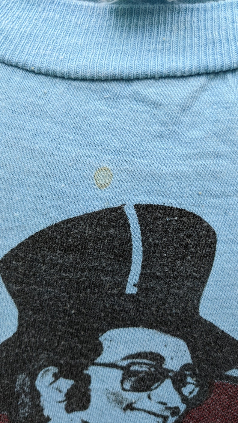 K&ouml;nnte beinhalten: Ein hellblaues T-Shirt mit einem Schwarzwei&szlig;bild einer Person mit Hut und Sonnenbrille. Das Bild ist leicht verblasst.