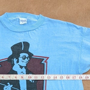 K&ouml;nnte beinhalten: Ein hellblaues T-Shirt mit einem Aufdruck eines Mannes mit Zylinder und Stock. Der Aufdruck ist in Schwarz, Wei&szlig; und Rot gehalten. Das Shirt hat einen gerippten Rundhalsausschnitt.