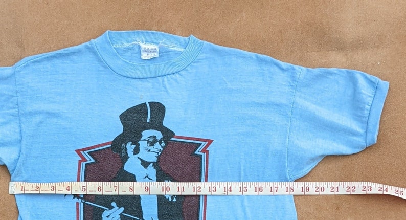 K&ouml;nnte beinhalten: Ein hellblaues T-Shirt mit einem Aufdruck eines Mannes mit Zylinder und Stock. Der Aufdruck ist in Schwarz, Wei&szlig; und Rot gehalten. Das Shirt hat einen gerippten Rundhalsausschnitt.