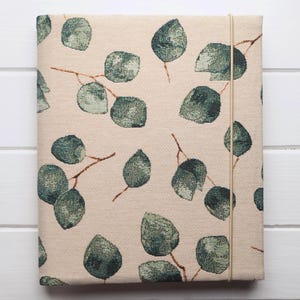 Peut inclure: Une couverture de journal en tissu de couleur crème avec un motif répétitif de feuilles d'eucalyptus vertes. La couverture est dotée d'une fermeture élastique de couleur crème.