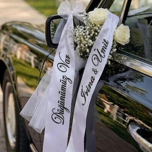 Könnte beinhalten: Ein schwarzes Hochzeitsauto, dekoriert mit weißen Bändern, Blumen und Text. Auf den Bändern stehen die Namen "Irina & Umit" und anderer Text. Weiße Rosen und Schleierkraut sind auf dem Auto angeordnet.