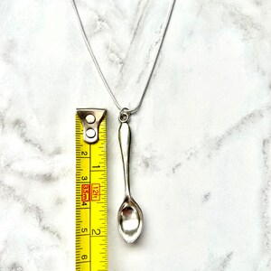 May include: A silver spoon pendant necklace on a silver chain. The spoon is about 2 inches long and is measured against a yellow tape measure.