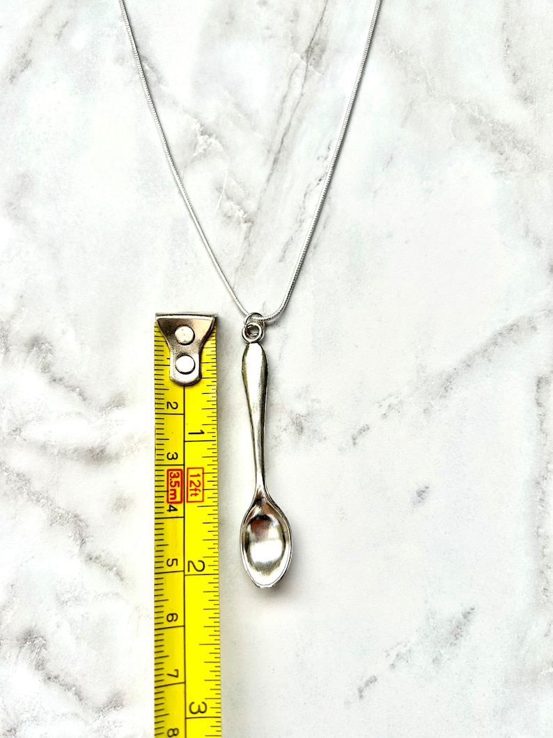 May include: A silver spoon pendant necklace on a silver chain. The spoon is about 2 inches long and is measured against a yellow tape measure.