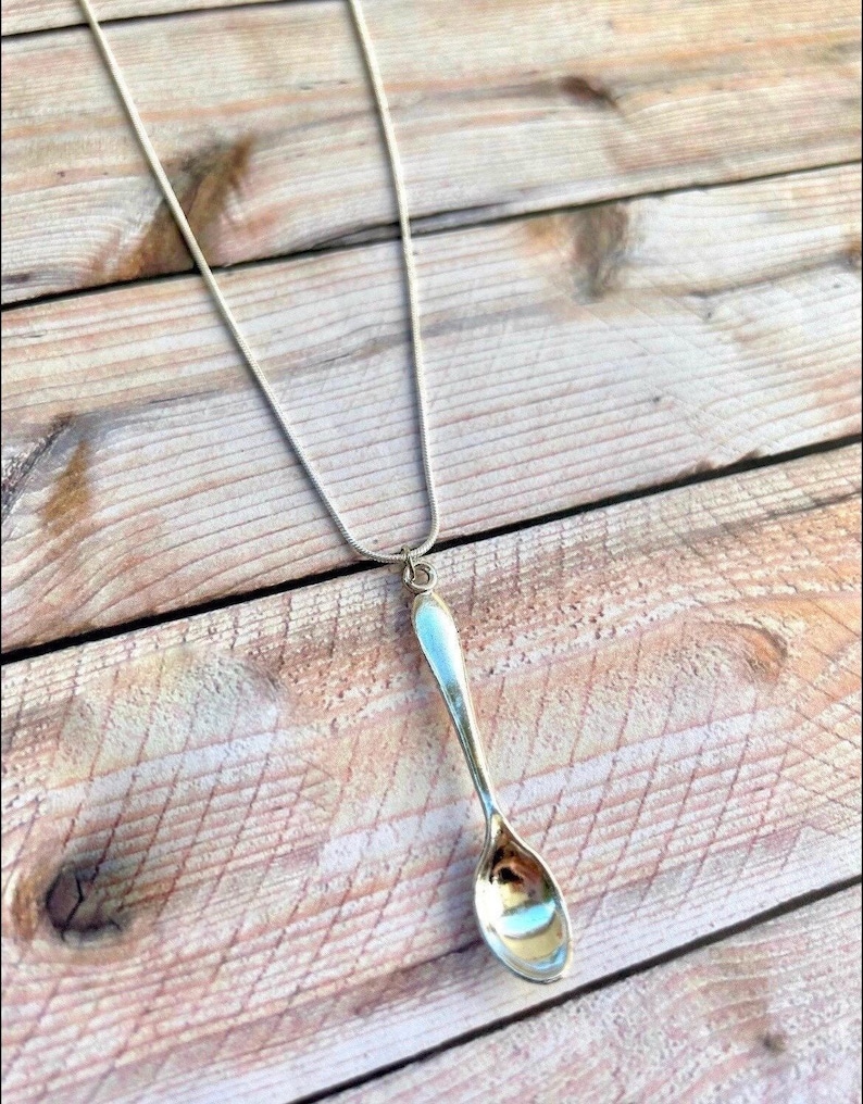 May include: A silver spoon pendant necklace on a silver chain. The spoon is hanging from the chain and is facing down. The necklace is on a wooden surface.