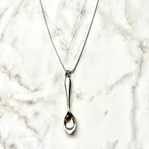 May include: A silver spoon pendant necklace on a silver chain. The spoon is hanging from the chain and is the focal point of the necklace.