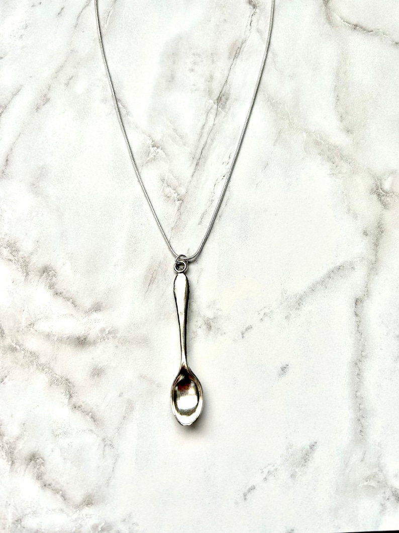 May include: A silver spoon pendant necklace on a silver chain. The spoon is hanging from the chain and is the focal point of the necklace.
