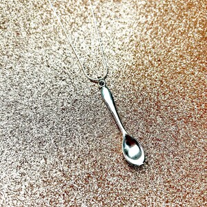 May include: A silver spoon pendant necklace on a silver chain. The necklace is on a sparkly gold background.