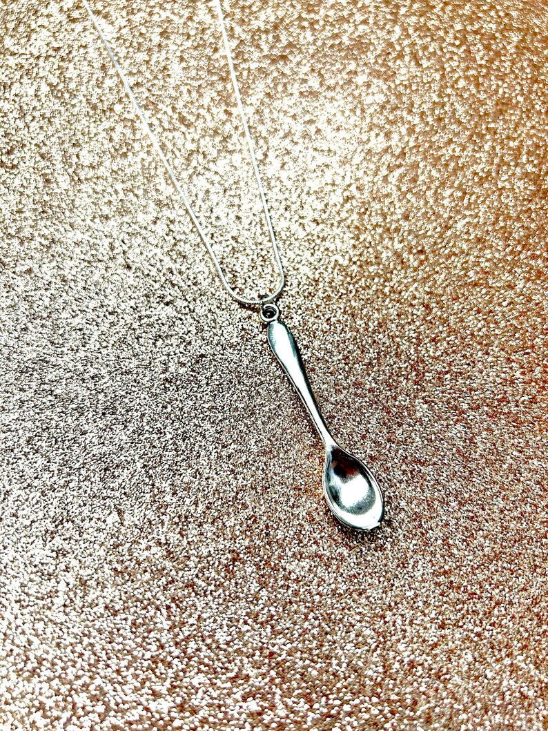May include: A silver spoon pendant necklace on a silver chain. The necklace is on a sparkly gold background.