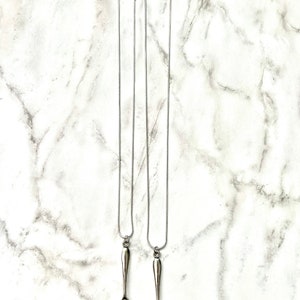 May include: Two silver spoon necklaces on a white marble background. The spoons are attached to silver chains.