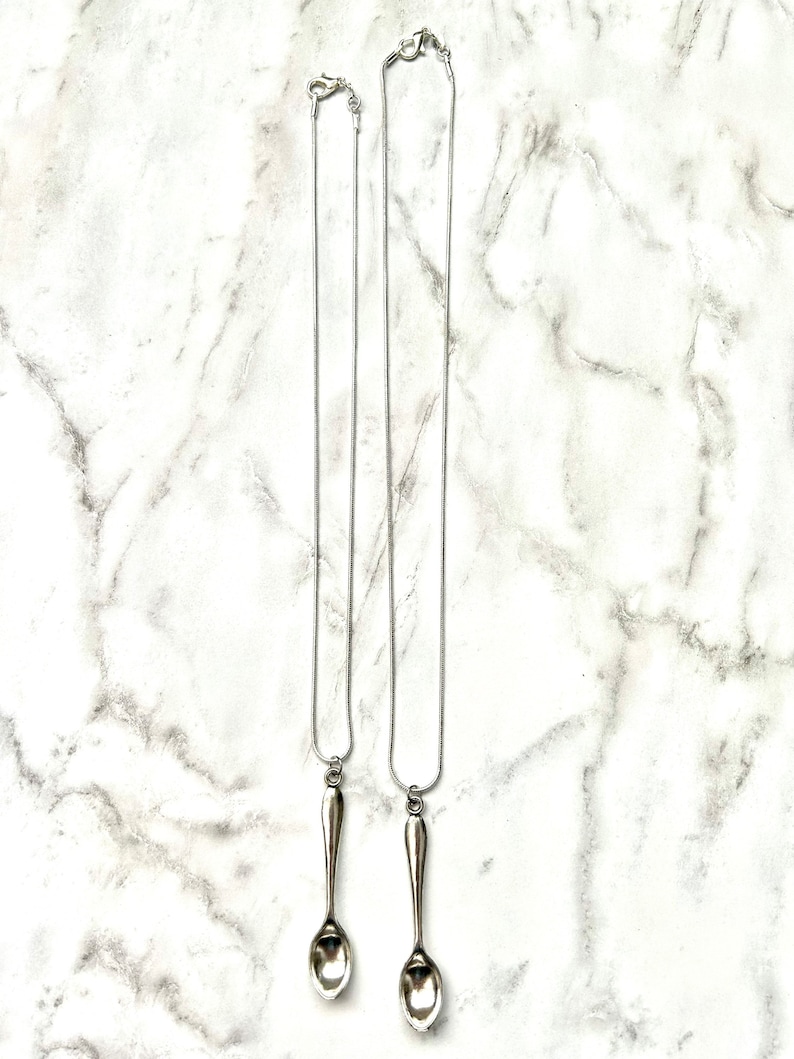 May include: Two silver spoon necklaces on a white marble background. The spoons are attached to silver chains.