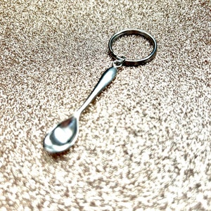以下が含まれることがあります： リングが付いた銀のスプーンのキーホルダー。スプーンは小さく、丸いボウルが付いています。キーホルダーはキラキラした金の背景の上にあります。