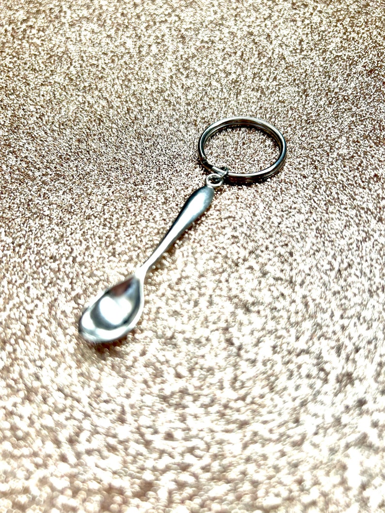 以下が含まれることがあります： リングが付いた銀のスプーンのキーホルダー。スプーンは小さく、丸いボウルが付いています。キーホルダーはキラキラした金の背景の上にあります。