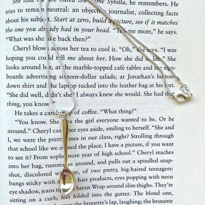 May include: A silver teaspoon with a rounded handle and a reflective surface. The spoon is resting on a page of a book with text about a woman named Sybilla.