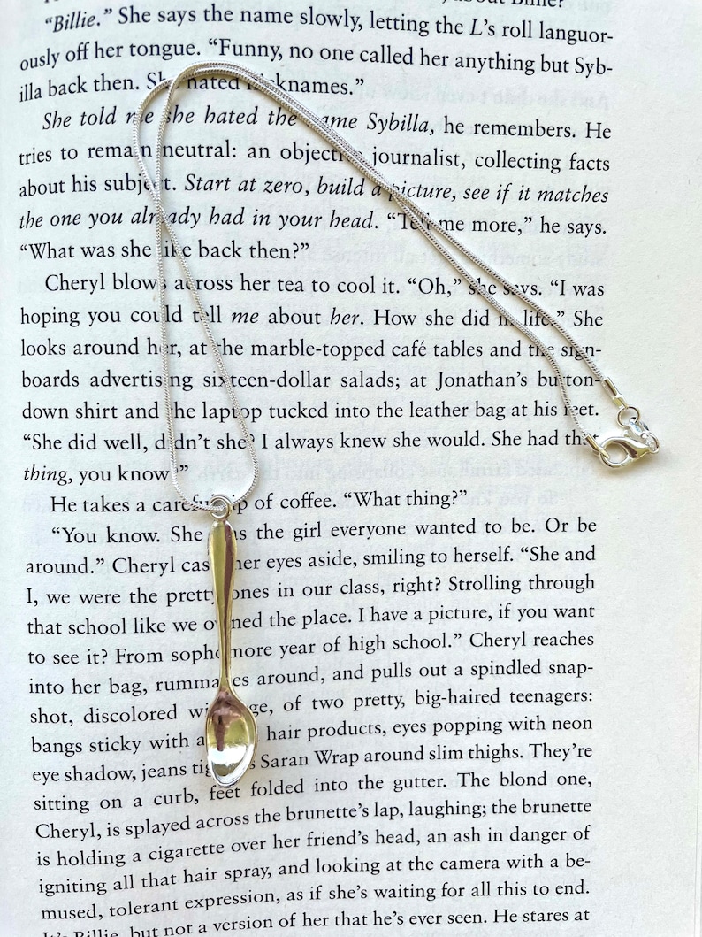 May include: A silver teaspoon with a rounded handle and a reflective surface. The spoon is resting on a page of a book with text about a woman named Sybilla.