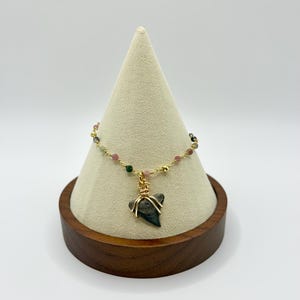May include: A gold-colored chain bracelet with small, colorful beads and a shark tooth pendant. The shark tooth is gray and has gold wire wrapped around it. The bracelet is displayed on a beige cone-shaped stand with a wooden base.